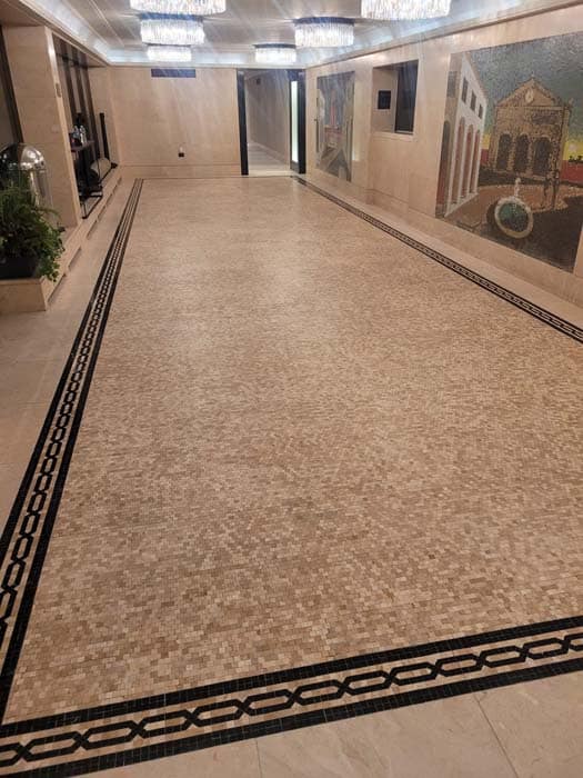 A long, empty hallway with a light mosaic tile floor bordered by a black geometric pattern reveals its elegance. The walls feature two large, colorful mosaic artworks depicting architectural scenes. A plant sits in a metal stand on the left side, embodying marble restoration from Astoria, NY.
