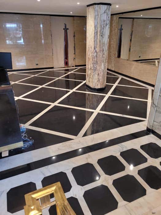 Luxurious room with glossy black and white geometric marble flooring, a large central column with a marbled design, mirrored walls, and a partially visible desk with a gold chair in the foreground. Perfect for those seeking top-notch marble restoration in Astoria NY.