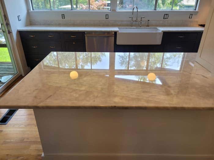 A kitchen with a large marble island countertop, dark lower cabinets, a farmhouse sink, and windows providing natural light. The countertop reflects two pendant lights and the outdoor scenery, showcasing the beauty of expert marble restoration in Astoria NY.