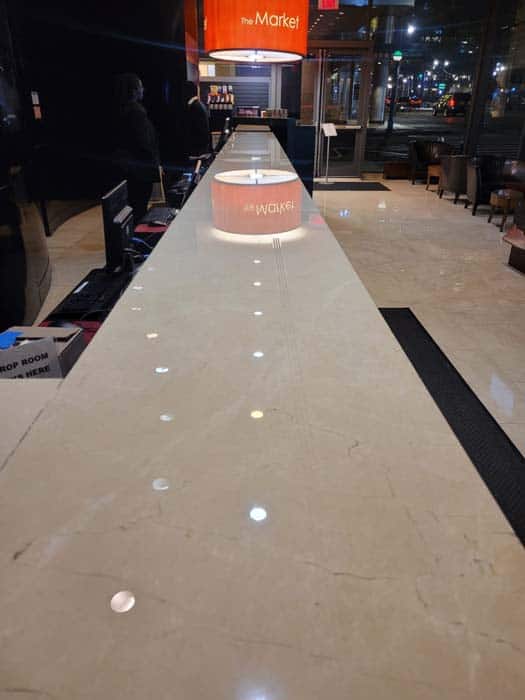 Long, sleek hotel reception counter with a shiny marble surface. A large, orange sign at the end reads "The Market." LED lights illuminate the countertop, showcasing flawless marble restoration Astoria NY. People stand near the entrance in the background.