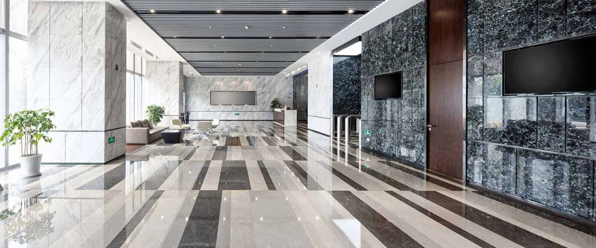 Spacious modern lobby with glossy striped marble floors and walls, benefiting from recent marble restoration Astoria NY. Two flat-screen TVs are mounted on a dark marble wall. Large windows usher in natural light, while several chairs and potted plants complete the decor.