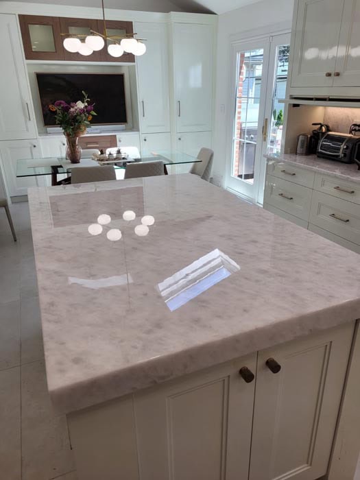 A modern kitchen features a large marble island countertop, showcasing the artistry typical of marble restoration in Astoria, NY. A dining area with a glass table and chairs sits in the background, adorned with flowers. Bright windows and white cabinets frame the space, with a toaster oven on the counter.