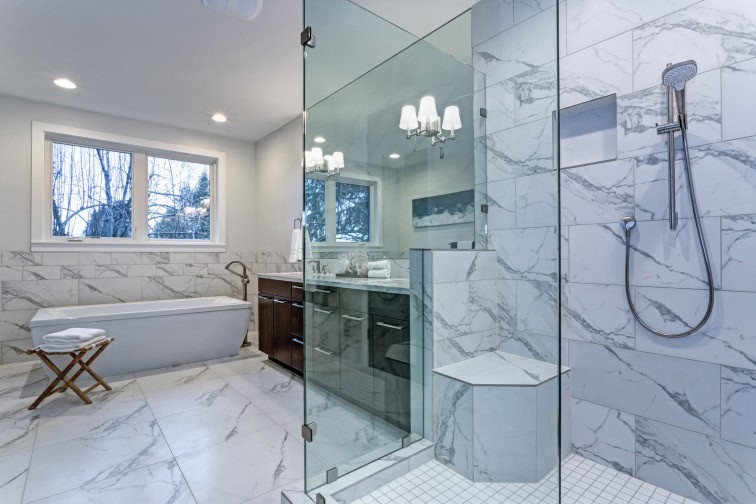 Modern bathroom with marble tile design. Features a glass shower, freestanding bathtub, wooden stool, and vanity with lit mirror. Recently completed marble restoration in Astoria, NY enhances the elegance. A window offers a view of trees outside, enhancing natural light.