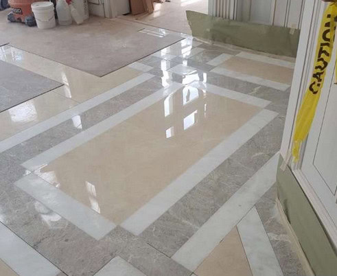 A polished floor featuring a geometric pattern of beige, gray, and white tiles gleams brilliantly after marble restoration in Astoria NY. Reflections dance on the shiny surface, while construction materials and caution tape suggest ongoing work in the background.