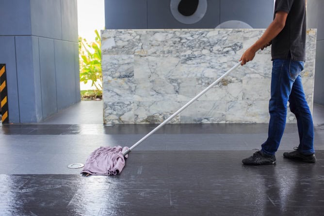 A person cleaning a tiled floor with a mop in what seems to be a lobby, possibly post-marble restoration Astoria NY. They are dressed in jeans and black shoes. A marble wall and part of a plant accentuate the indoor setting.