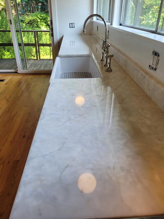 A long kitchen counter with a polished marble surface, recently enhanced by marble restoration in Astoria NY, features a deep sink and modern faucet. It overlooks a wooden deck through a large glass door, where sunlight streams through windows to illuminate the countertop and wooden floor.