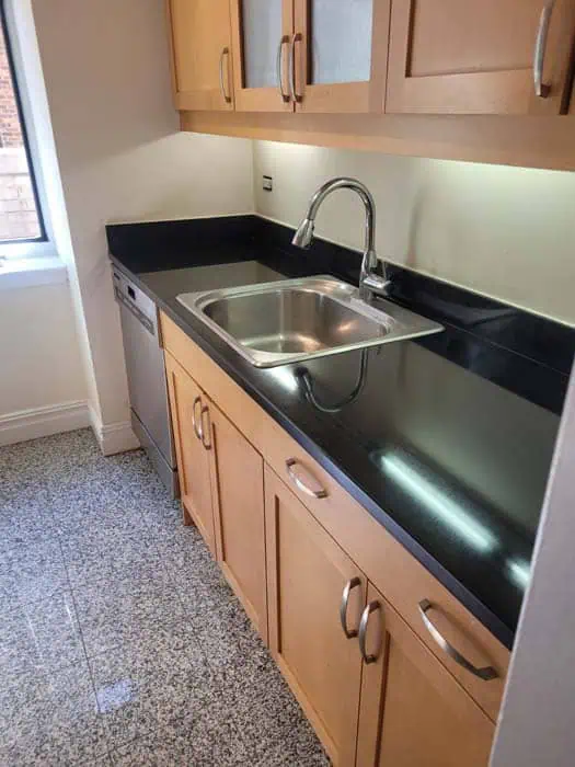 A compact kitchen with wooden cabinets, a stainless steel sink, black countertops, a dishwasher, and a window on the left. The floor boasts speckled gray tiles reminiscent of sleek stone restoration Manhattan NY. LED lights illuminate the countertop area.