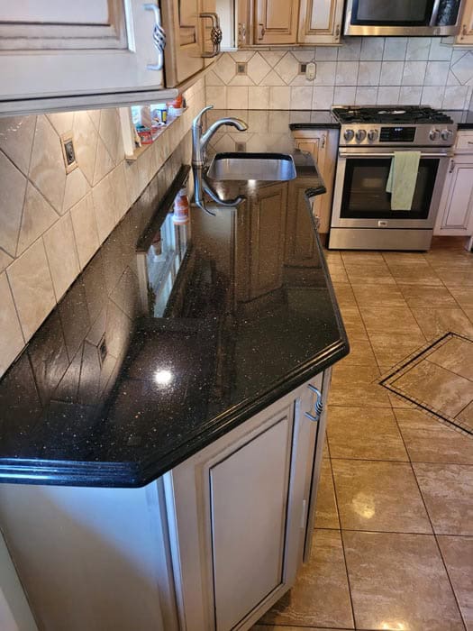 A kitchen with beige cabinets, black granite countertops, and a stainless steel double sink. The backsplash features tan tiles. A gas stove with an oven and a range hood is in the background. Thanks to marble restoration in Astoria NY, the light brown tiled floor gleams like new.