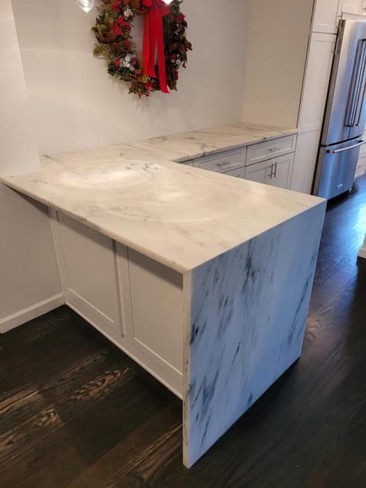A modern kitchen boasts white marble countertops with subtle gray veining, reflecting recent marble restoration in Astoria NY. The L-shaped counter pairs perfectly with sleek white cabinetry and a stainless steel refrigerator, while a floral wreath adds charm to the wall.