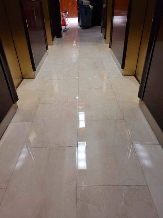 A polished marble hallway with reflective flooring showcases the finesse of marble restoration in Astoria, NY. The corridor is flanked by dark elevator doors, while at the end, a cleaning cart and trash bin are visible. Bright lighting casts vivid reflections on the floor.