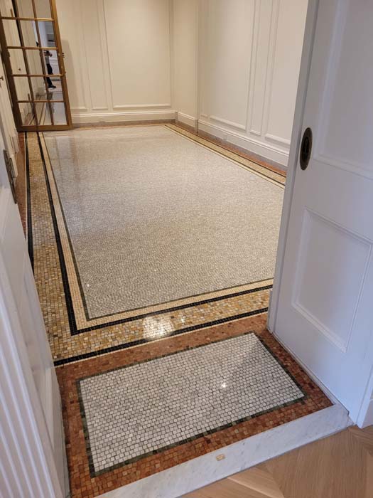 A room with beige walls features a detailed mosaic tile floor, bordered by black, white, and brown tiles similar to those seen in marble restoration projects in Astoria, NY. A wooden door partially opens into the space, revealing reflections on the glossy surface.