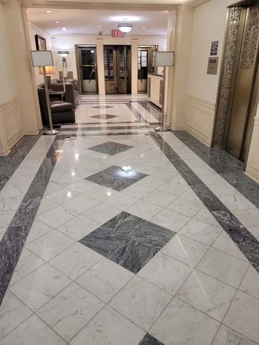 A hallway with a shiny, patterned tile floor leads to a lobby area. The lobby features a sofa, lamps, decorative columns, and an elevator on the right. Fresh from marble restoration in Astoria NY, beige walls and ceiling lighting create a warm atmosphere. A fire exit sign is visible above.