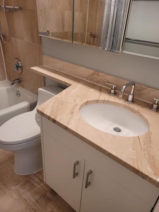 Bathroom with a beige marble countertop, single oval sink, silver faucet, and white cabinet. Wall-mounted medicine cabinet above. Bathtub with showerhead and beige tiled walls in the background showcases expert marble restoration Astoria NY craftsmanship.