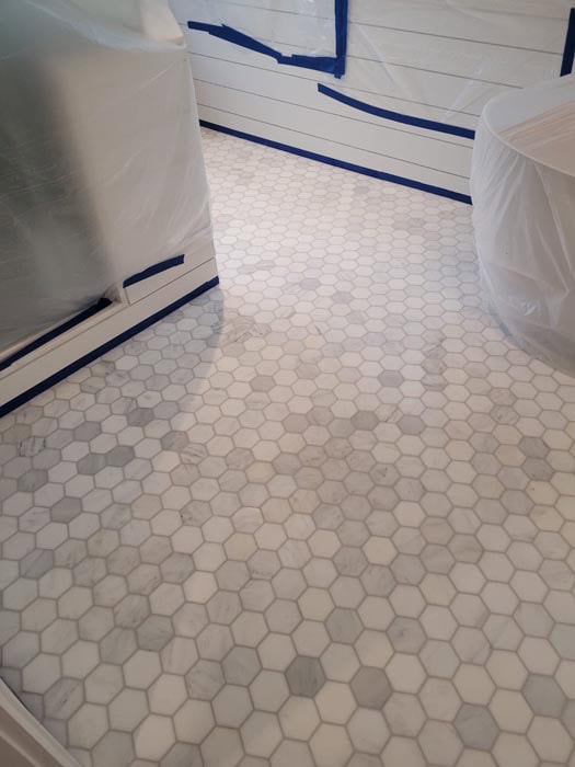 Hexagonal white and light gray tiles cover the floor of a room under renovation, echoing marble restoration techniques from Astoria, NY. Furniture is wrapped in plastic, and the walls are shielded with taped sheets. Light streams through an unseen window, illuminating the tiled pattern.