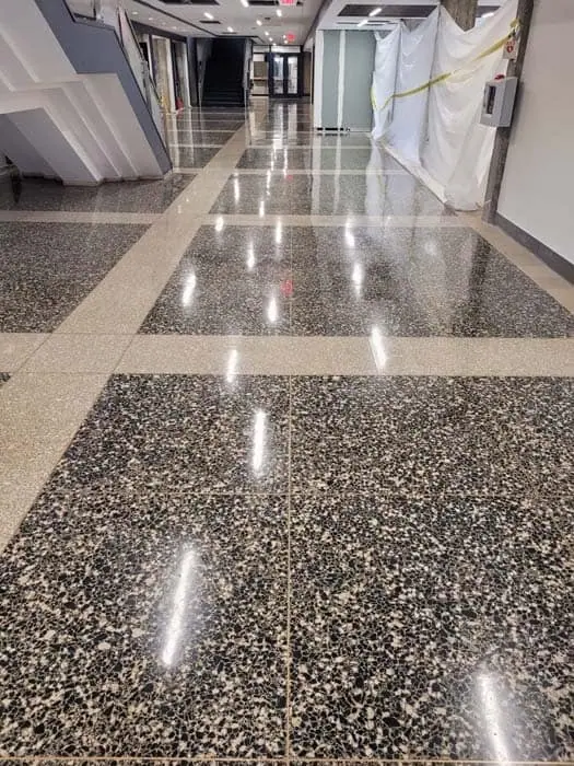 A shiny polished floor in a hallway with reflective surfaces showcases the beauty of marble restoration in Astoria, NY. The left side is bordered by a staircase, and the right side has a wall partially covered with plastic sheets. Bright overhead lights reflect on the floor's surface.