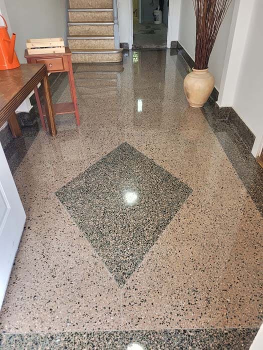 A hallway with polished terrazzo flooring features a large diamond pattern in the center, showcasing expert stone restoration. A wooden table and chair sit to the left, while a tall vase with twigs stands on the right, and stairs ascend gracefully in the background.