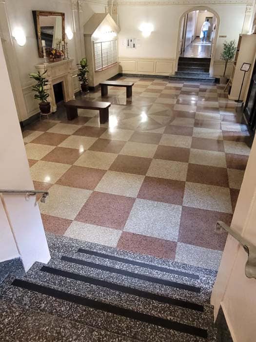 A spacious lobby with a polished checkered floor in beige and brown hues showcases expert marble restoration, reflecting elegance. Two wooden benches sit in the center, as potted plants flank a decorative fireplace. The stairs descend into the lobby, leading to a hallway with an arched entrance.