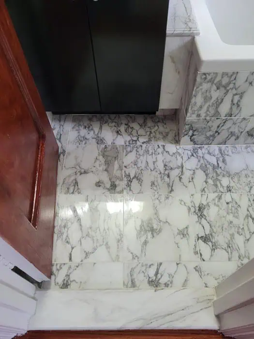 Marble-tiled bathroom floor with gray and white patterns, partially bordered by a wooden door on the left. A dark cabinet and a corner of a white bathtub are visible at the top, showcasing an ideal setting for expert marble restoration in Astoria, NY.