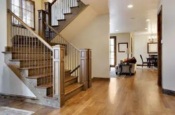 A warm, spacious hallway with polished hardwood floors and a wooden staircase adorned with metal railings on the left showcases exquisite marble restoration, while soft lighting illuminates the area. A cozy living space with a couch and dining table invites you in further.