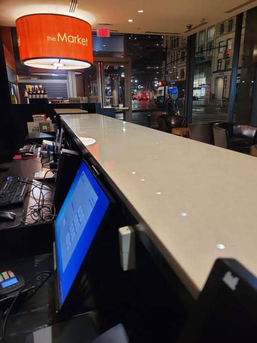 The hotel reception area at night showcases a long counter, reminiscent of the elegance brought by marble restoration in Astoria, NY. The illuminated "The Market" sign and computer screens stand out against the city street view visible through the windows.
