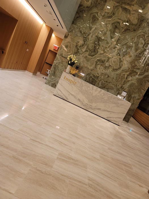 A modern lobby interior with a sleek marble reception desk, a marble-patterned wall, and light-colored tiled floors showcases the results of expert marble restoration in Astoria, NY. A bouquet of flowers graces the counter. The decor is minimalist and sophisticated.