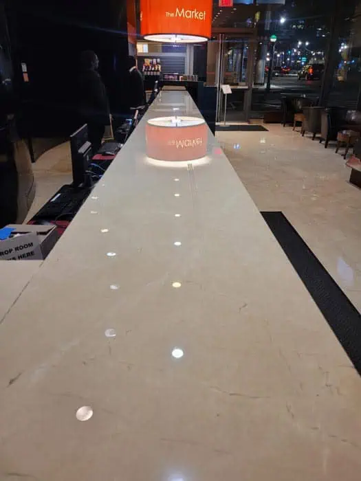 A long, polished marble countertop in a modern interior space gleams after expert stone restoration Manhattan NY, catching reflections of ceiling lights. A sign in the background reads "The Market." There are chairs and tables to the side, and large windows reveal city lights outside.