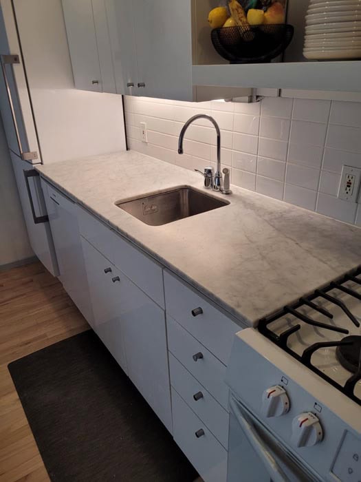 Modern kitchen with white cabinets and a gleaming marble countertop, enhanced by a stainless steel sink. A fruit bowl and dishes grace the shelf above. A gas stove sits on the right, complemented by under-cabinet lighting and hardwood flooring. Perfect spot for marble restoration Astoria NY enthusiasts.