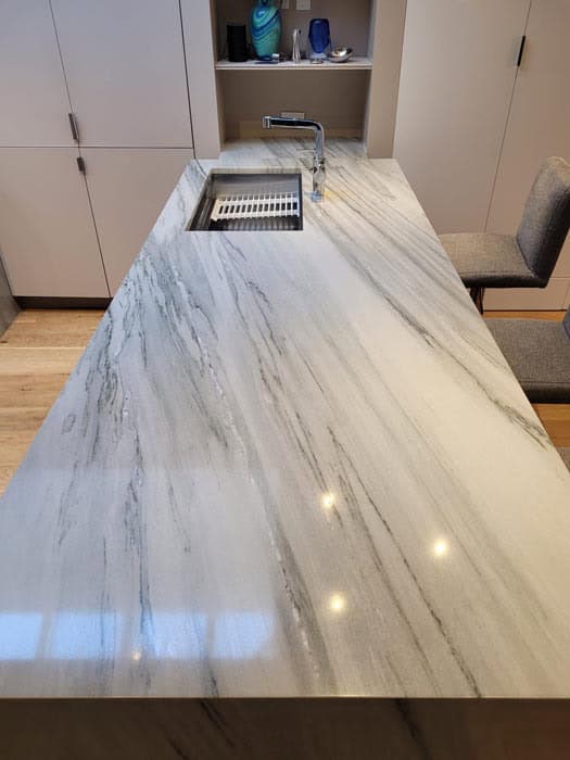 A modern kitchen with a long, marble island countertop featuring grey veining, reminiscent of expert stone restoration Manhattan NY. It includes a built-in sink and faucet. In the background, there are shelves with various items and a cushioned chair on the side.