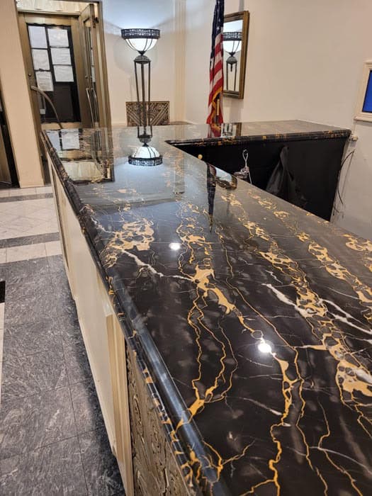 A sleek reception area featuring a polished black marble counter with striking gold veining, expertly maintained by marble restoration in Astoria, NY. Behind the counter is a mirror-adorned wall with a decorative lamp and a U.S. flag in the corner. The floor boasts an elegant mix of black and white tiles.