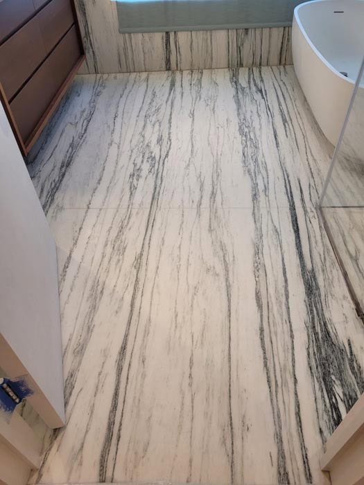Bathroom with a large white and gray marble floor, expertly maintained with marble restoration Astoria NY services. A wooden cabinet sits on the left, while a pristine white freestanding bathtub graces the right, next to a glass shower wall that's partially visible.
