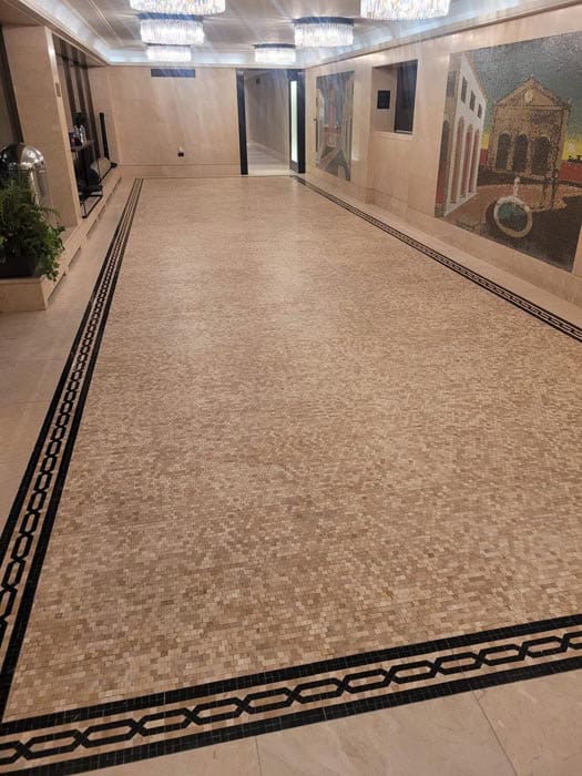 A spacious hallway with a light-colored mosaic tile floor, beautifully restored to its original charm, is bordered by a black pattern. Walls feature abstract artwork, and chandeliers hang from the ceiling, illuminating the corridor with warm light.