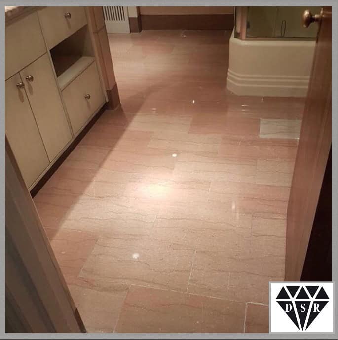 A polished marble floor in a bathroom with beige cabinets on the left and a bathtub in the background. The smooth, shiny surface reflects light, showcasing expert marble restoration Astoria NY craftsmanship. A logo with "DSR" inside a diamond shape is in the bottom right corner.