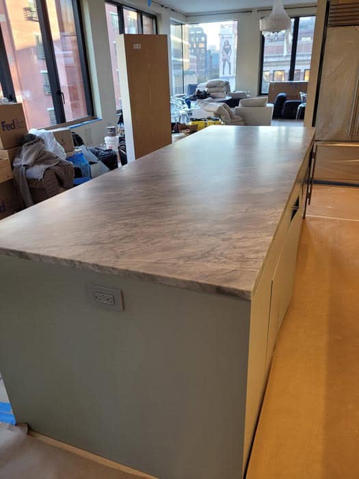 A modern kitchen boasts a large marble countertop island with electrical outlets. The room, mid-renovation, is filled with construction materials and boxes. Large windows reveal a city view, hinting at the need for future stone restoration in Manhattan NY to maintain its pristine look.