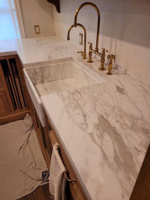 A kitchen with a white marble countertop and matching sink, exemplifying the elegance found in stone restoration Manhattan NY. Brass fixtures, including a tall faucet and handles, complement the decor. Below, a beige towel hangs on a rod with a loose cloth nearby.