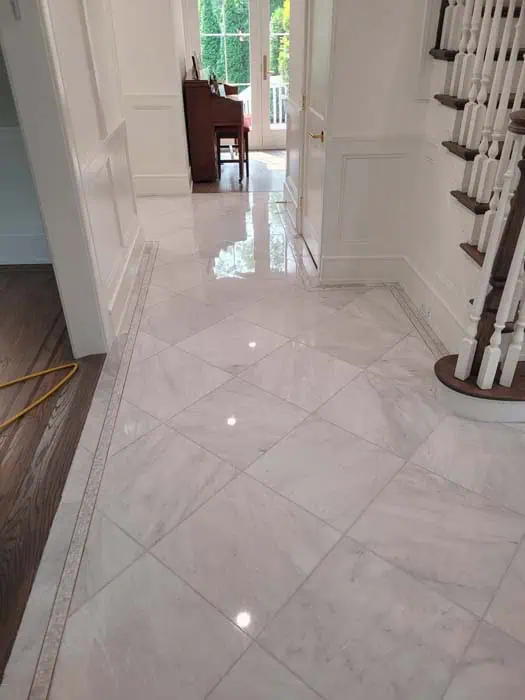 A hallway with glossy white marble tiles, possibly the result of expert marble restoration in Astoria NY, reflects light from a nearby window. A wooden staircase with white spindles is on the right. Natural light streams through an open door leading to a balcony or patio, with a piano visible in the background.