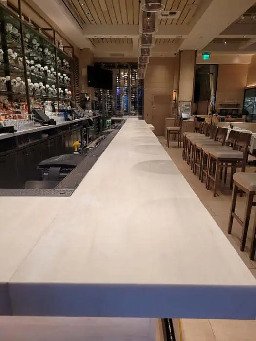 An empty bar with a sleek, long white countertop and tall chairs. Shelves stocked with bottles illuminate the background, complemented by modern overhead lighting. This well-lit space exudes a contemporary charm akin to high-end marble restoration in Astoria, NY.