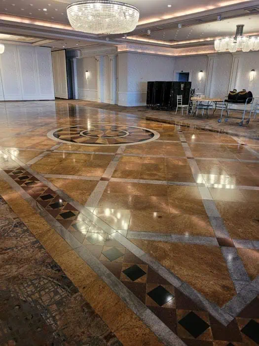 Spacious ballroom with ornate marble floor featuring geometric patterns, recently rejuvenated by experts in marble restoration Astoria NY. Several large chandeliers hang from the ceiling. A folded table and cart are visible in the background near white-paneled walls.