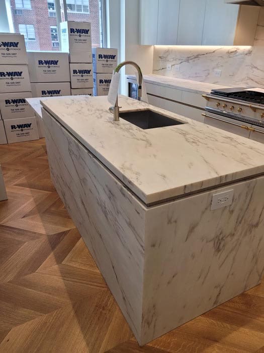A modern kitchen with a marble island featuring an integrated sink and sleek faucet. The wooden herringbone floor complements the marble surfaces. Boxes labeled "R-WAY Moving" are stacked near the window, hinting that marble restoration in Astoria NY might soon be on the agenda.