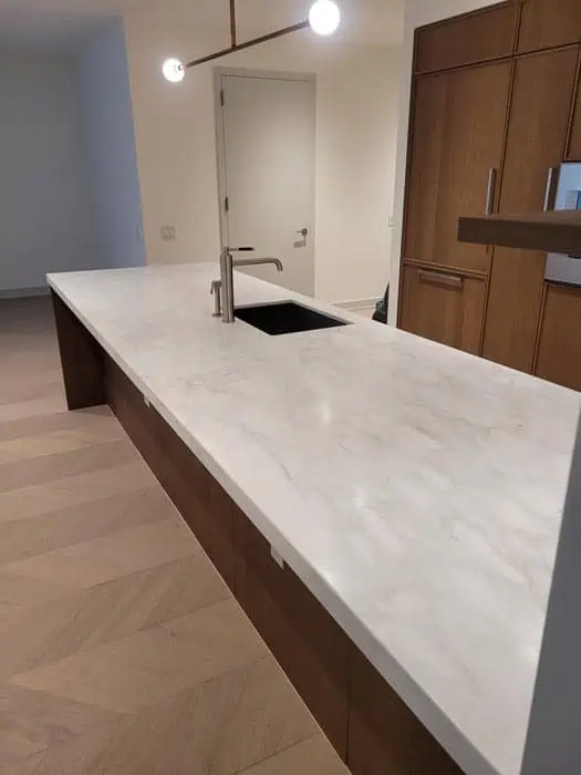Spacious modern kitchen featuring a long white marble countertop, expertly maintained with marble restoration in Astoria, NY. A built-in black sink and faucet complement the design. Wooden cabinets line the wall, while a sleek light fixture hangs above, all atop a herringbone wood-patterned floor.