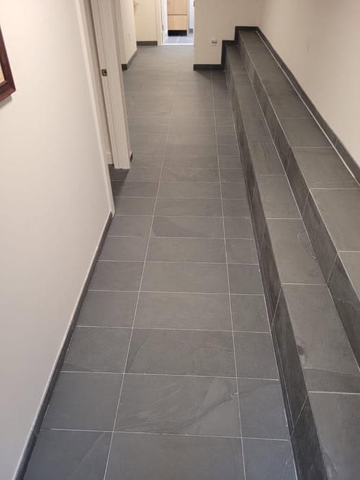 A hallway with gray tiles on the floor and an adjacent three-tiered step area on the right. The white walls showcase a sleek design, while a small section of wood is visible at the distant end, echoing elegance fit for any marble restoration project in Astoria, NY.