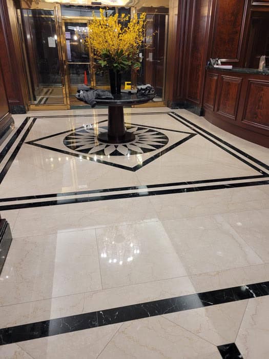 Elegant hotel lobby with glossy marble floor featuring black geometric patterns, expertly maintained with marble restoration Astoria NY. A central round table displays a large arrangement of vibrant yellow flowers. Dark wooden panels and glass entrance doors enhance the luxurious atmosphere.