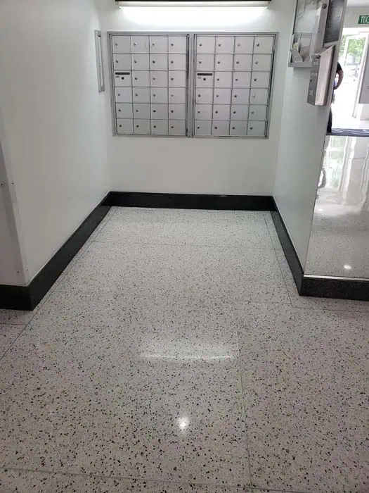 A hallway with white walls and a speckled floor, reminiscent of marble restoration in Astoria NY, leads to a set of mailboxes mounted on the back wall. The area is lit by overhead lights, and a door with a glass panel is visible on the right side.