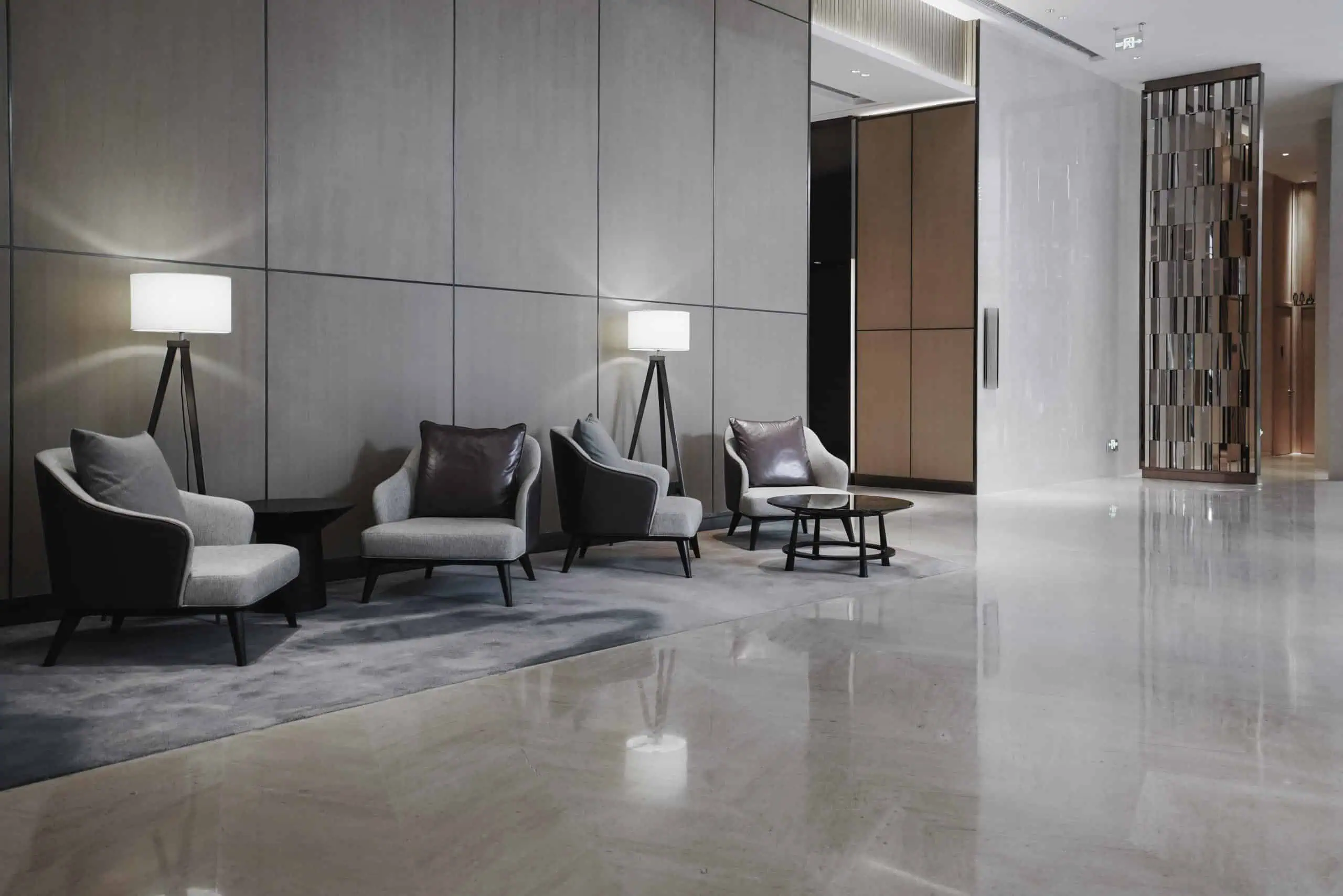 Modern lobby area with three cushioned chairs and two standing lamps on a large gray rug. The space, boasting marble restoration from Astoria, NY, features sleek wooden panels, marble flooring, and a decorative partition. The atmosphere is minimalistic and contemporary.