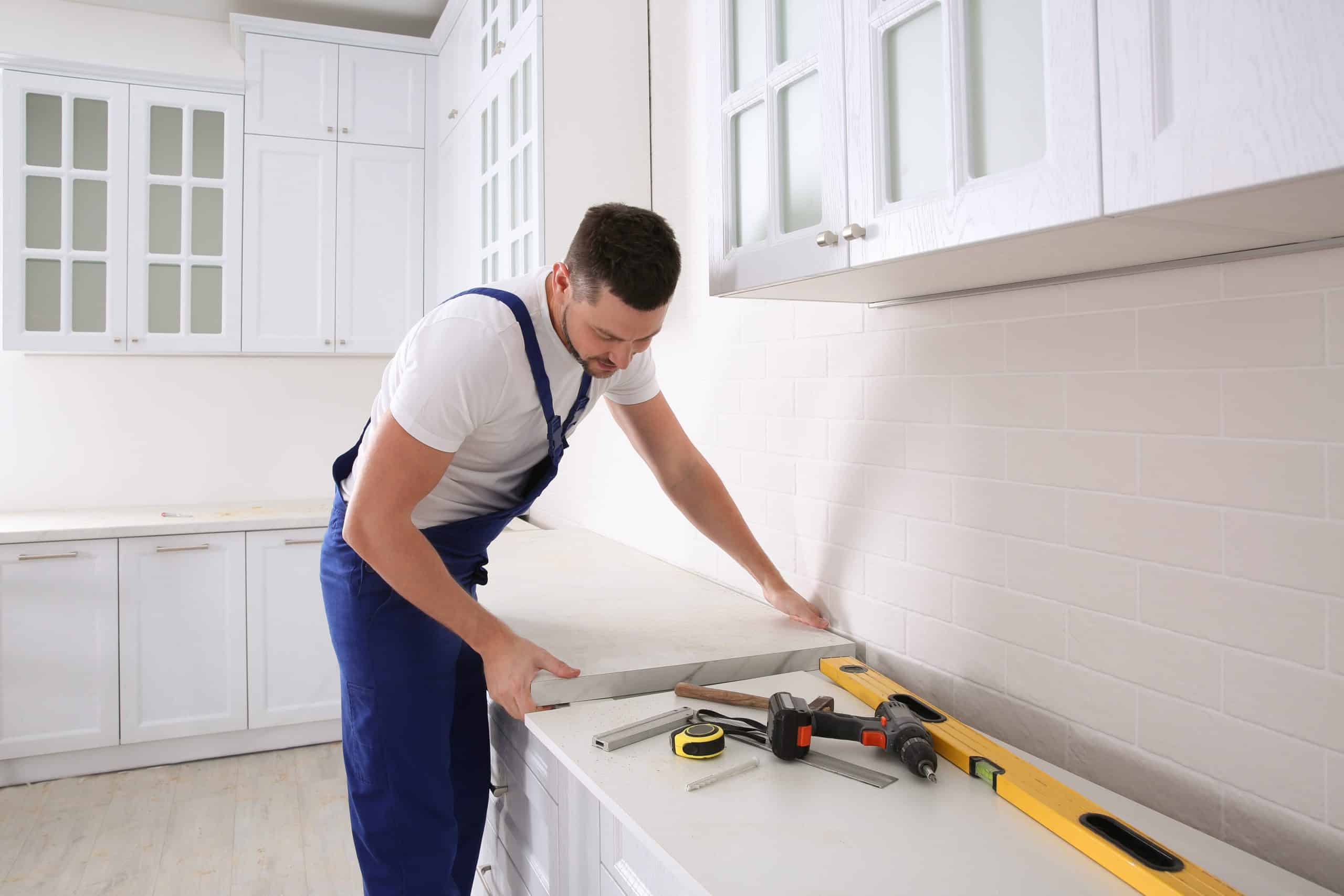A man in a white T-shirt and blue overalls is installing a kitchen countertop, showcasing skills akin to marble restoration in Astoria, NY. He’s placing the countertop on white cabinets, among various tools like a spirit level and hammer. The kitchen features white cabinets and light-colored tiles.