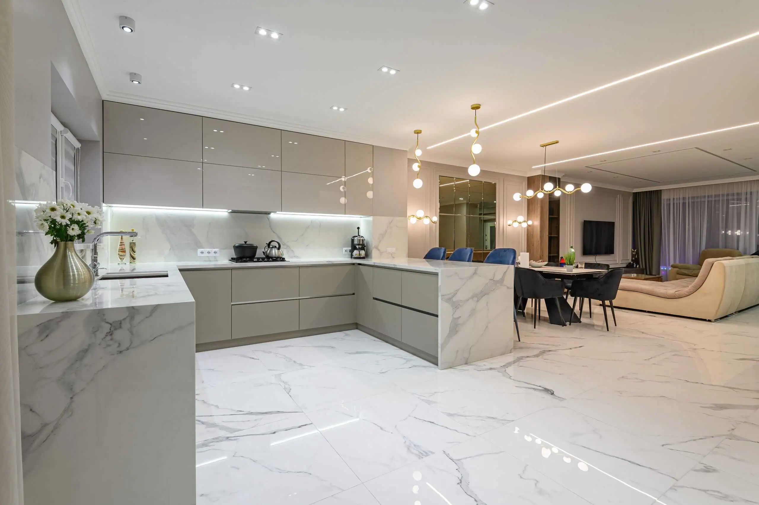 Modern kitchen and dining area with expertly restored marble flooring and countertops. Beige cabinets, stainless steel appliances, and a gold vase with flowers adorn the kitchen. Dining table with blue chairs and pendant lights; beige couch in adjoining living room reflects marble restoration elegance.