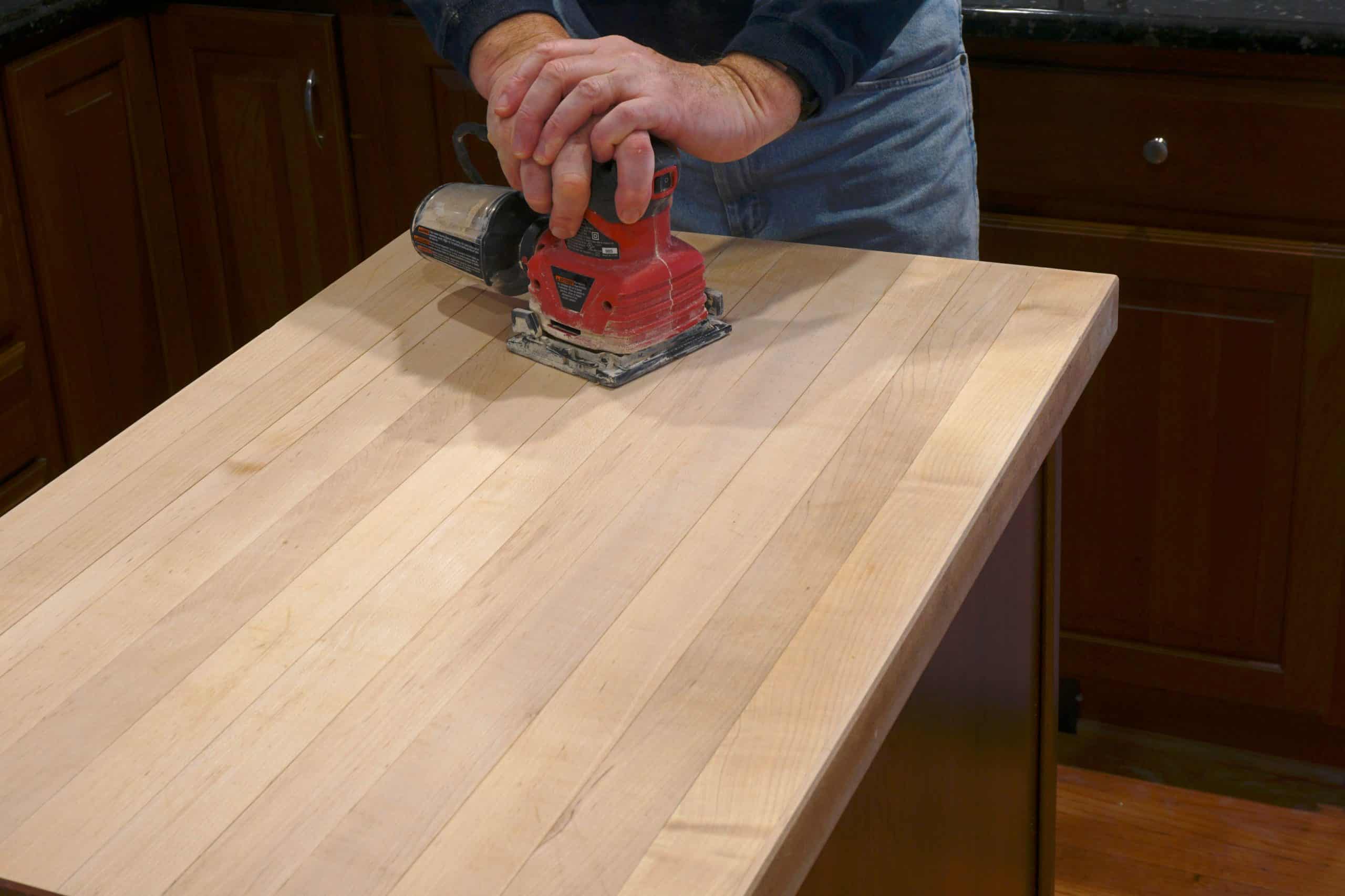 A person is sanding a wooden countertop with a red electric sander in a kitchen setting, hinting at the meticulous attention usually seen in marble restoration Astoria NY. The countertop boasts a light wood finish, with wooden cabinets enhancing the backdrop.