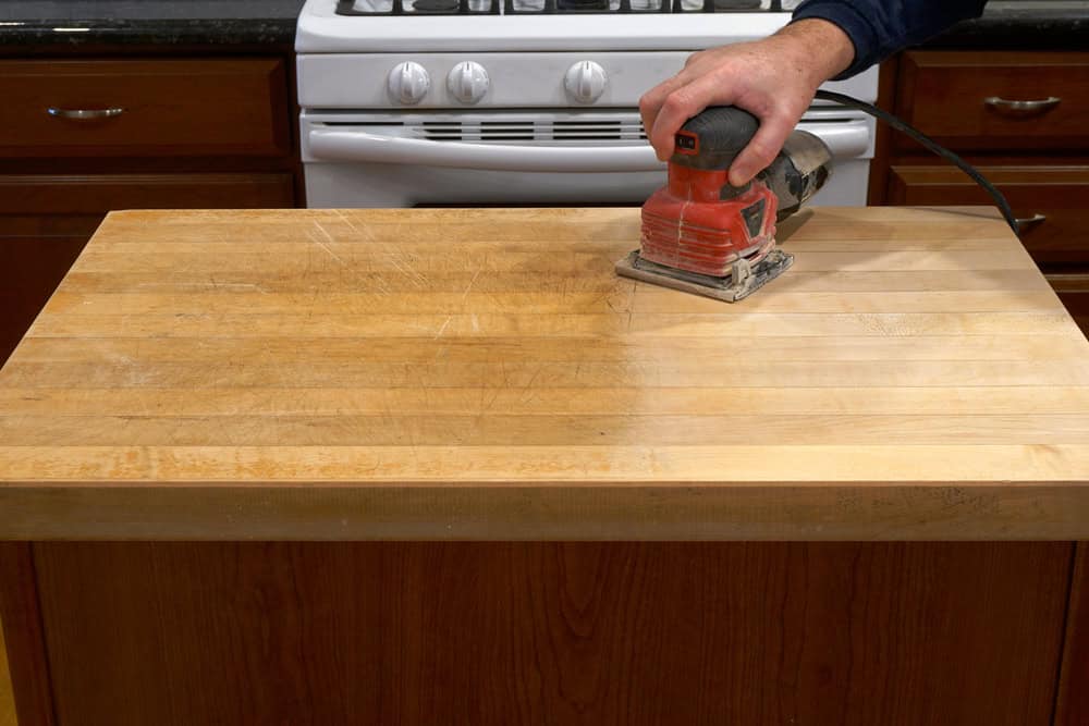 A hand uses a red electric sander on a wooden kitchen island, smoothing its surface. The island is partially sanded, showing a contrast between the sanded and unsanded wood. With skills akin to marble restoration Astoria NY experts, precision transforms this space. A stove is visible in the background.