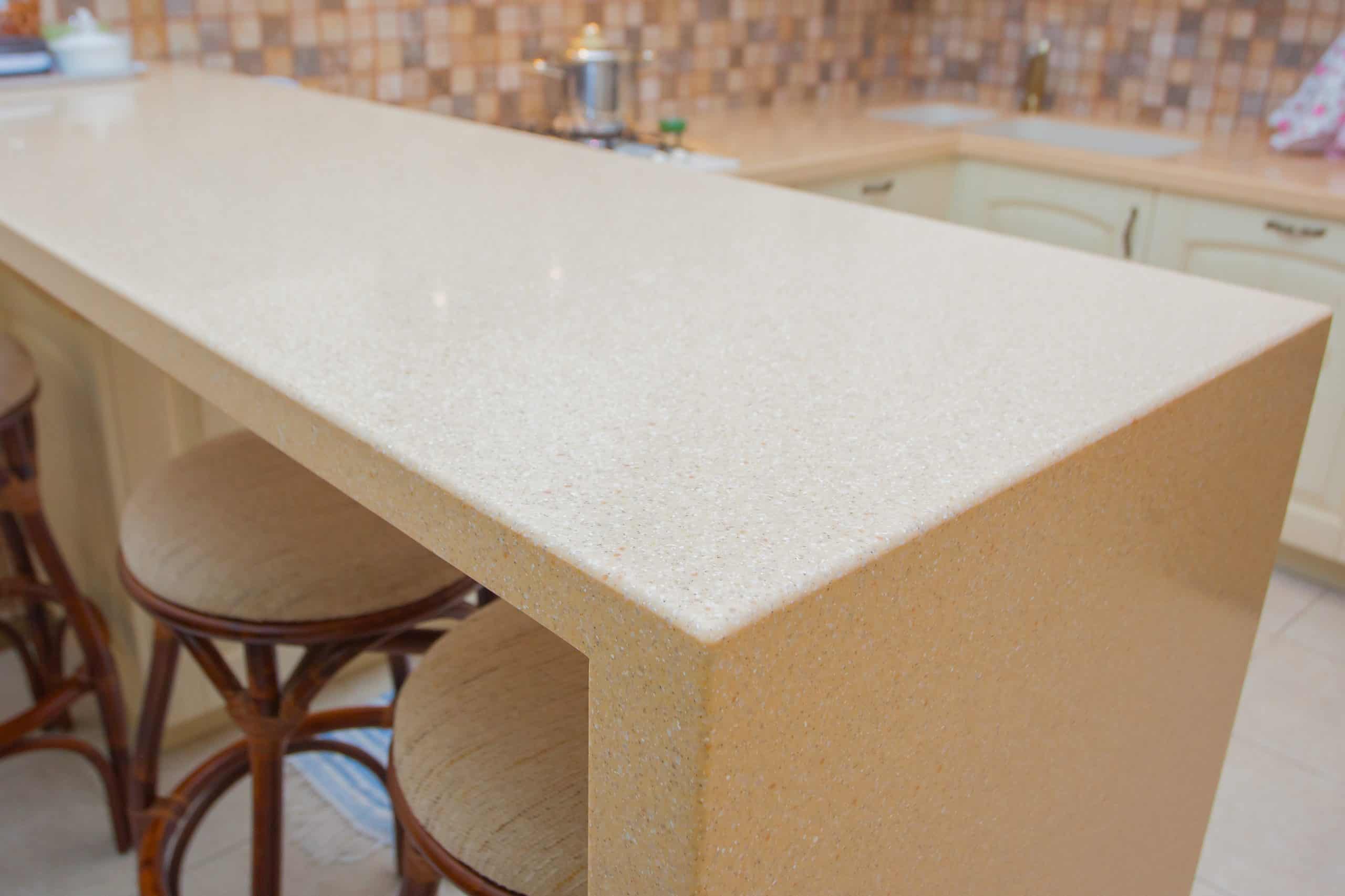 A modern kitchen with a smooth, beige countertop featuring stone restoration Manhattan NY expertise. Three wooden stools with cushioned seats are tucked underneath. The background showcases a tiled backsplash alongside part of a stove with a pot and sink basin.