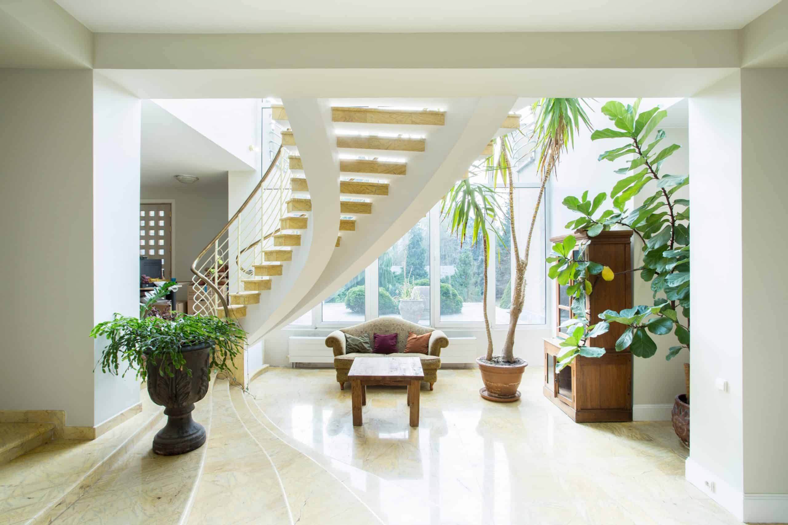 A bright living space featuring a spiral staircase, large windows, and indoor plants. An elegant sofa and wooden coffee table rest on a shiny marble floor, enhanced by expert marble restoration Astoria NY services. The room is filled with natural light.