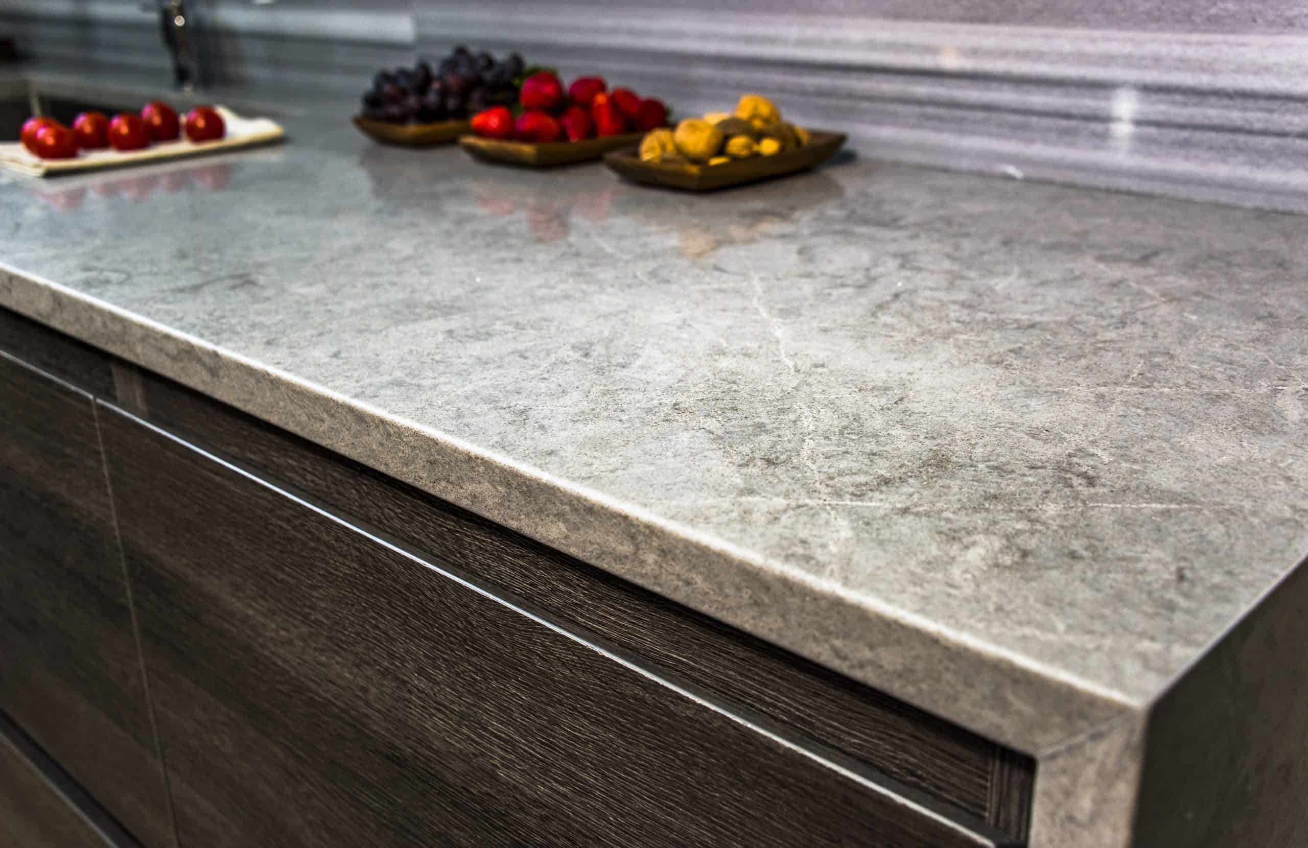 Marble kitchen countertop with a gray, slightly reflective surface. In the background, plates hold cherry tomatoes, grapes, raspberries, and nuts. This stunning piece exemplifies expert marble restoration from Astoria, NY, seamlessly set atop a dark wood cabinet.